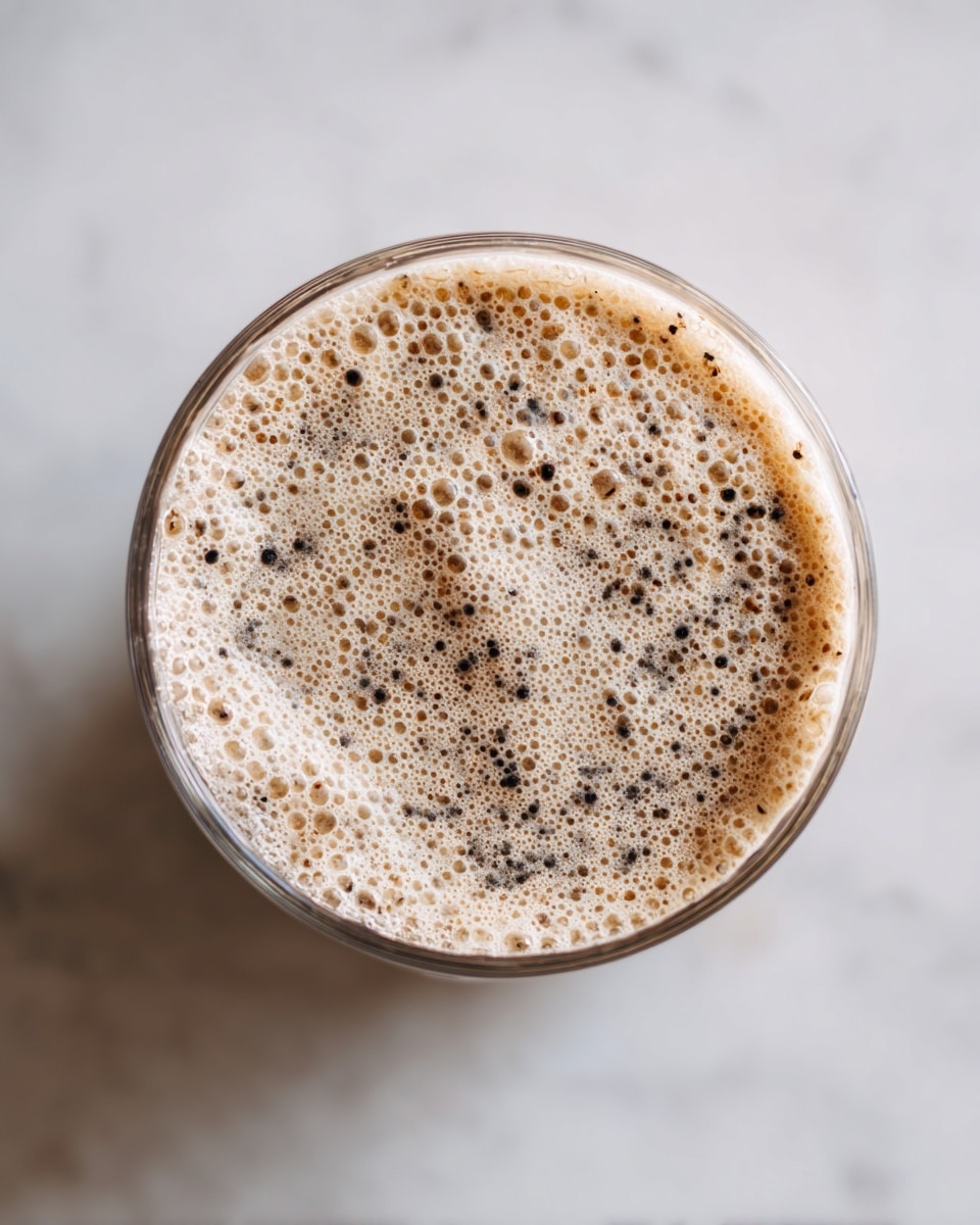 The image shows a close-up top view of a clear blender cup filled with a light beige foam layer that has many small bubbles on the surface. Scattered black and dark brown specks are mixed into the foam, giving it a textured look. The blender cup is placed on a white marbled surface with a clean and bright background. The focus is on the creamy, bubbly layer inside the cup, showing a smooth and airy texture with tiny floating particles. photo taken with an iphone --ar 4:5 --v 7