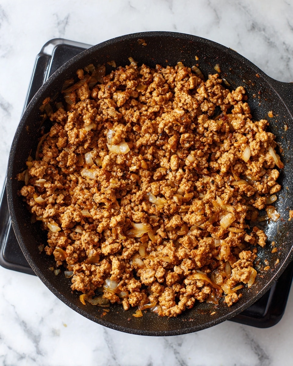 A black cast iron pan filled with cooked ground meat mixed with small pieces of onions, showing a brown and slightly golden color. The pan is placed on a black stove burner with a white marbled background visible behind and around the stove. The ground meat has a crumbly texture with some bits clumped together, and there is some oil or moisture giving a slight shine to the mixture. Photo taken with an iphone --ar 4:5 --v 7