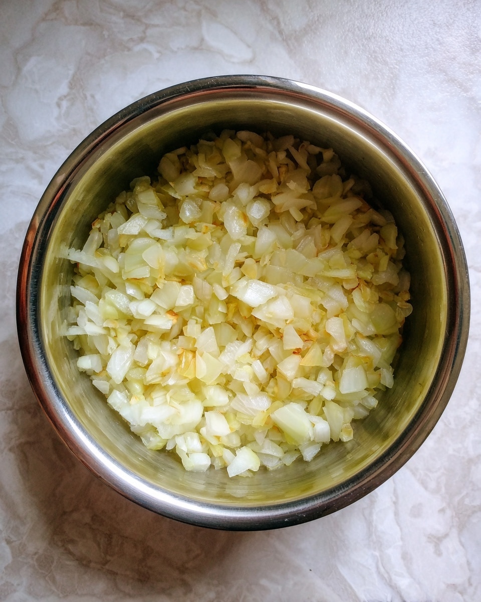 A shiny silver pot filled with one layer of small, chopped onions that are soft and slightly yellow from cooking. The onions are spread evenly across the bottom of the pot, which has a reflective surface showing hints of light. The background is a white marbled texture. photo taken with an iphone --ar 4:5 --v 7