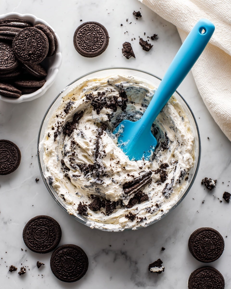 A clear glass bowl filled with thick white cream mixed with broken pieces of dark chocolate sandwich cookies, showing black cookie crumbs and white cream bits scattered through the mixture. Inside the bowl, a bright blue spatula is partly dipped into the creamy cookie mixture. Near the bowl, there is a white bowl filled with whole dark chocolate sandwich cookies, and several whole cookies are spread around on the white marbled surface. A soft white cloth is partially visible in the upper right corner. The whole scene is bright and clean with a white marbled background photo taken with an iphone --ar 4:5 --v 7