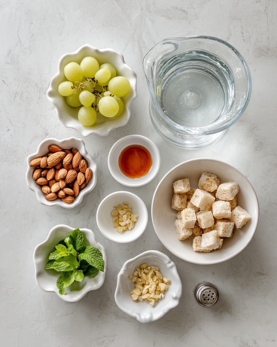 The image shows eight small white bowls and a glass pitcher arranged on a white marbled surface. Starting from the top right, there is a white bowl filled with small light beige bread cubes with a soft texture. Below it, there is a medium white bowl containing whole blanched almonds in a creamy light brown color. Below that, a small white bowl holds fresh bright green mint leaves. To the left, a small scalloped white bowl contains roasted almonds with a brown toasted surface. Above it, a tiny white bowl has pale yellow minced garlic with a soft texture. Slightly above and left, another small white bowl holds a reddish-brown liquid resembling a sauce. To the left of the bowl with almonds, a small white bowl contains several green grapes with smooth shiny skin. At the top left, there is a clear glass pitcher filled with water, showing the transparent liquid and the pitcher’s shape clearly. In the middle of the bowls, a small shiny silver salt shaker is visible. All elements sit neatly on the light grey marbled surface, creating a clean and fresh look. Photo taken with an iphone --ar 4:5 --v 7