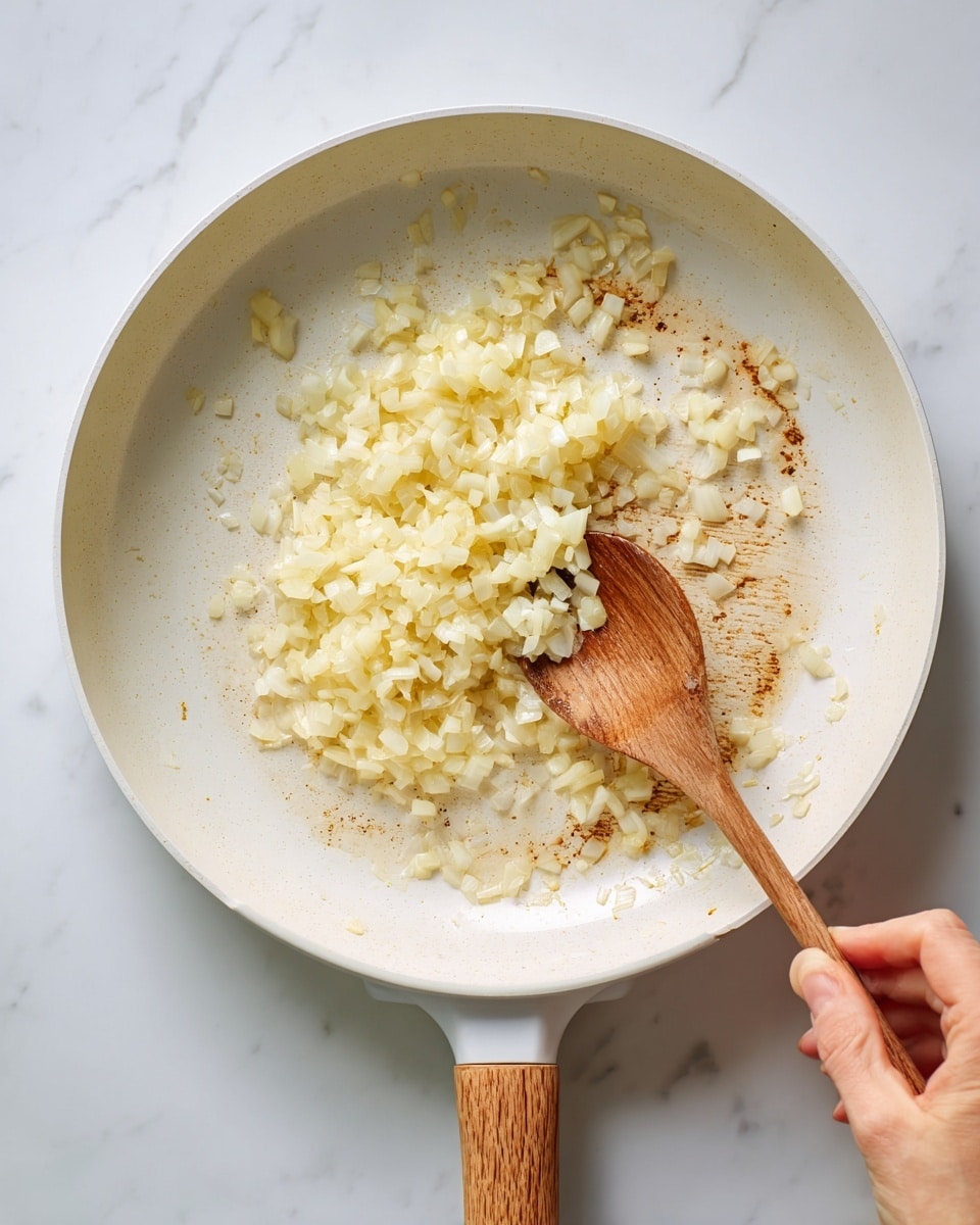 A white frying pan with a wooden handle sits on a white marbled surface. Inside the pan are small, soft-looking, pale yellow diced onions being stirred with a wooden spoon held by a woman's hand. The onions form a small pile mostly in the center of the pan, showing slight browning on the pan's surface around the edges. The overall scene is bright and clean, highlighting the cooking process. photo taken with an iphone --ar 4:5 --v 7