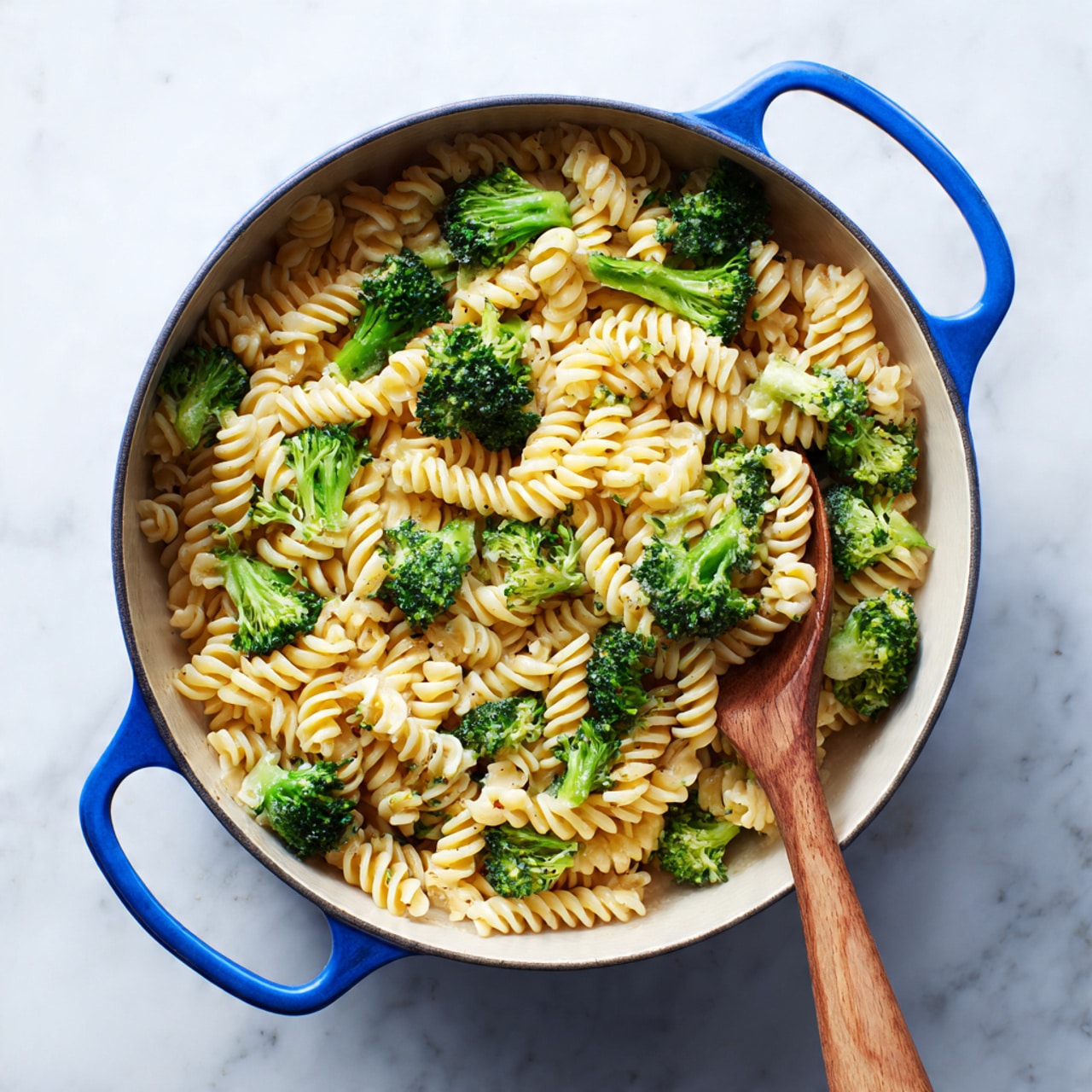 A large blue-handled pot filled with twisted spiral pasta mixed with bright green broccoli pieces, evenly spread and coating the pasta. A wooden spoon with a long handle rests inside the pot on the right side, partially covered with the pasta and broccoli mix. The pot is placed on a clean white marbled surface. Photo taken with an iphone --ar 4:5 --v 7