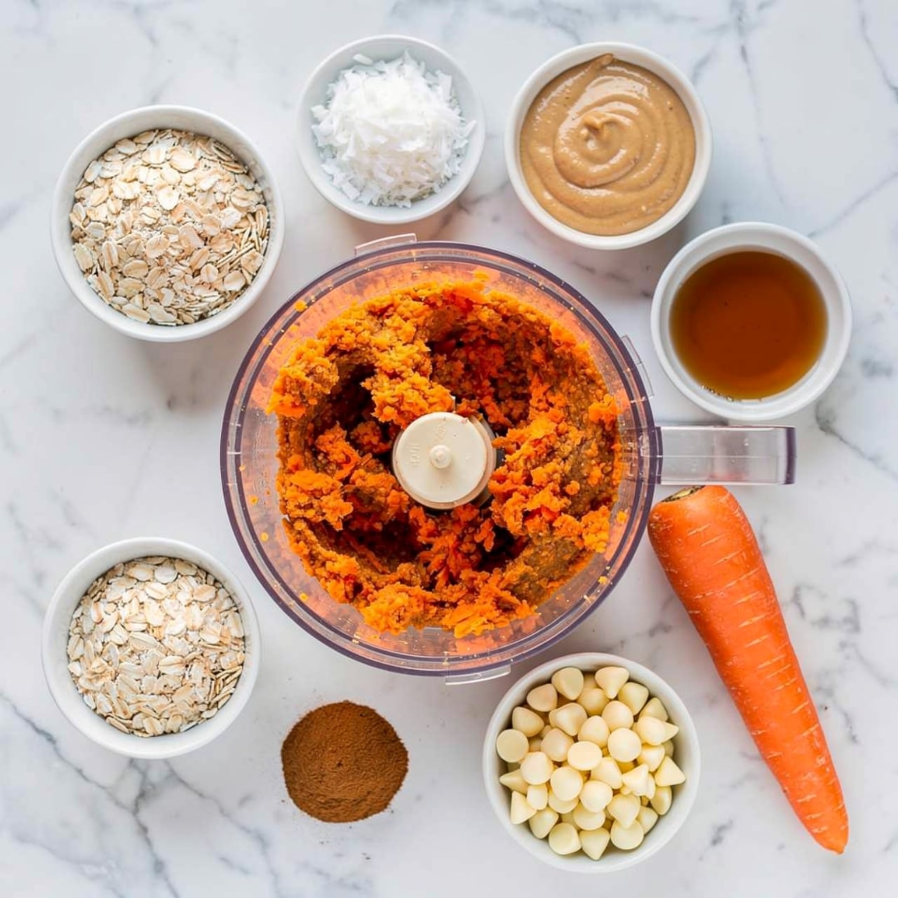 The image shows a clear food processor bowl filled with layers of different ingredients, sitting on a white marbled surface. The bottom layer appears to be beige rolled oats spread all around. On the left side, there is a bright orange layer of finely grated carrots. Above the oats and to the right, there is a thick, smooth, brown peanut butter layer with a small pool of dark amber liquid syrup on top. Near the center at the bottom, there are some pale sliced nuts. To the right of the nuts, there is a pile of medium brown powder (likely cinnamon), and a small patch of white salt is visible next to the powder. The plastic handle of the food processor bowl extends downwards in the front. Photo taken with an iphone --ar 4:5 --v 7