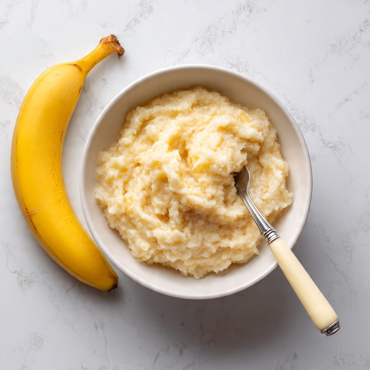 A white bowl holds a single layer of mashed banana with a chunky texture, its pale yellow color spread unevenly inside. A silver fork with a cream-colored handle rests in the bowl on the right side, partially covered by the mashed banana. Next to the bowl on the white marbled surface, two whole yellow bananas lie close together at the top left corner. The background is a smooth white marbled texture. photo taken with an iphone --ar 4:5 --v 7