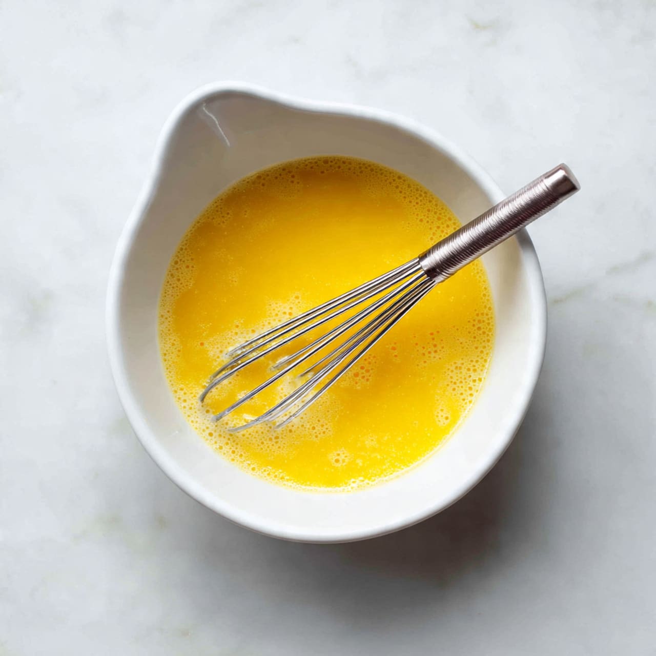 A white bowl with a small spout filled with a smooth, bright yellow mixture, likely beaten eggs, with a few small bubbles on the surface. A silver metal whisk rests inside the bowl, partially submerged in the liquid. The bowl is placed on a white marbled textured surface. photo taken with an iphone --ar 4:5 --v 7