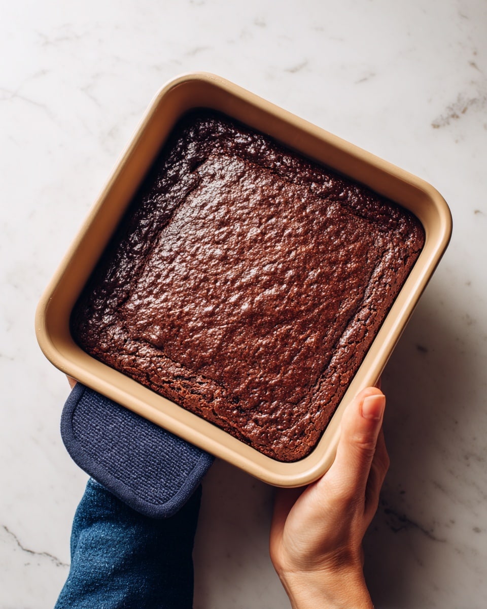 The image shows a freshly baked dark brown chocolate dessert, possibly brownies, in a rectangular light beige pan with smooth edges. The surface of the dessert is rough and uneven, showing small bumps and cracks all over. A woman's hand is holding the pan with a dark blue silicone oven mitt, gripping the edge firmly. The background is a white marbled surface. Photo taken with an iphone --ar 4:5 --v 7