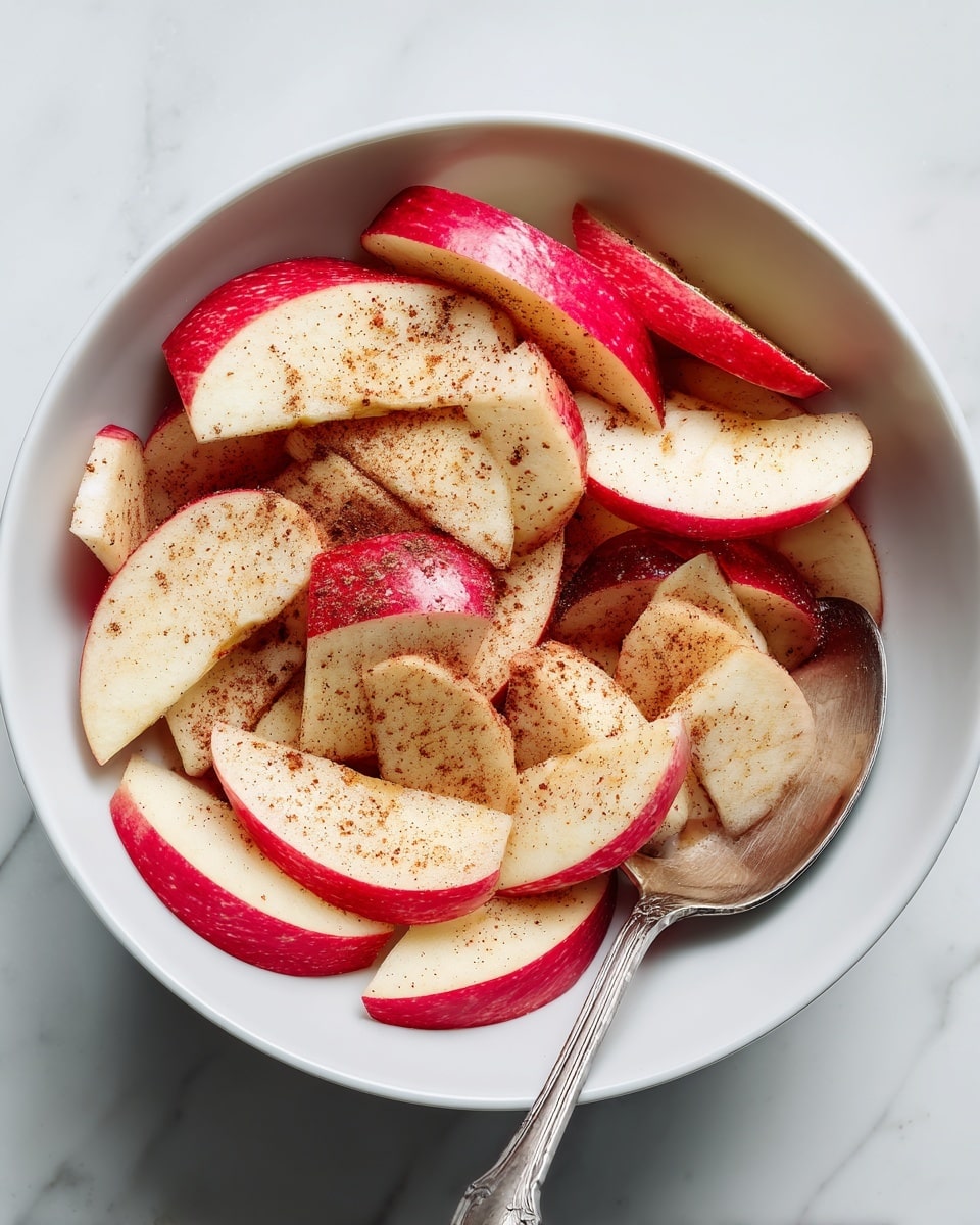 A white bowl filled with many thin apple slices, each slice showing red skin on one edge and pale yellow flesh on the rest. The slices are sprinkled with a light brown powder, giving them a dusty look. A shiny silver spoon rests on the right side inside the bowl, partially covered by apple slices. The bowl sits on a white marbled surface. photo taken with an iphone --ar 4:5 --v 7