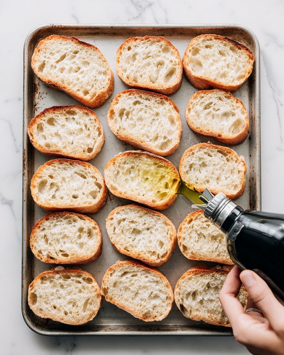 The image shows a tray with 16 toasted bread slices arranged in rows on a white marbled surface. Each slice has a golden brown crust with a soft, porous inside showing many small and medium holes, giving a light and airy texture. The tray is metal with a slightly worn look, and there is a black bottle with a silver spout held by a woman's hand near the bottom of the frame, likely pouring oil onto the bread. The scene is simple and bright with a clean background. photo taken with an iphone --ar 4:5 --v 7