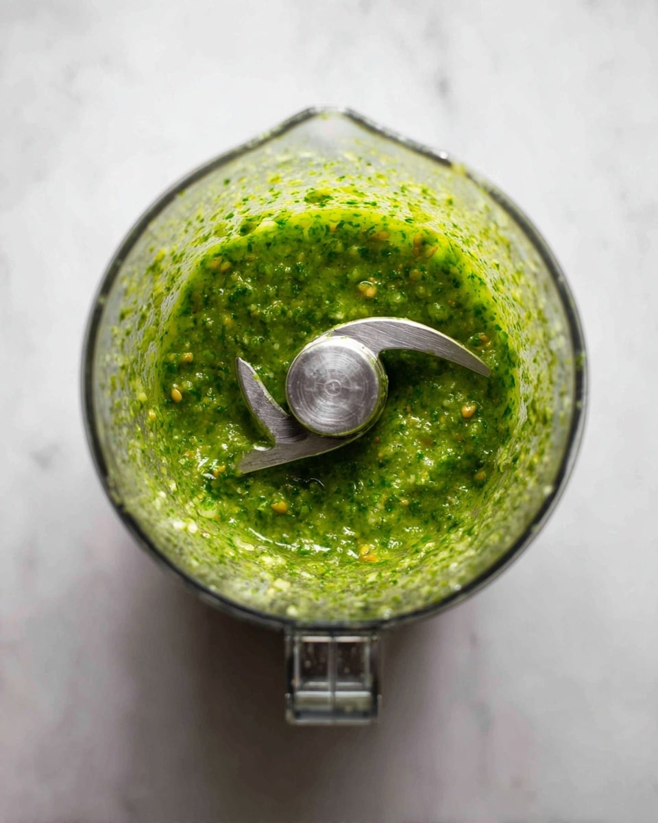 The image shows a clear blender jar viewed from the top, filled with a green sauce that has a slightly chunky texture with small darker green and yellow bits mixed inside. The blender blade is centered and visible in the middle, with two sharp silver blades curving toward opposite sides. The background and surface below the blender are a white marbled texture, creating a clean and bright setting. photo taken with an iphone --ar 4:5 --v 7