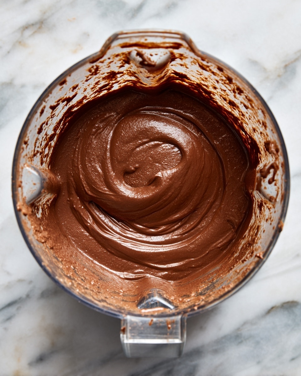 The image shows a close-up of a blender jar filled with thick dark brown sauce or paste. There is one main layer of the smooth, rich, thick sauce inside the blender, with bits of the mixture splattered and smeared around the clear jar’s inner walls. The blender jar is placed on a white marbled surface with some parts of the sauce visible outside the jar's edges. The lighting highlights the texture and shine of the sauce, making it look moist and dense. photo taken with an iphone --ar 4:5 --v 7