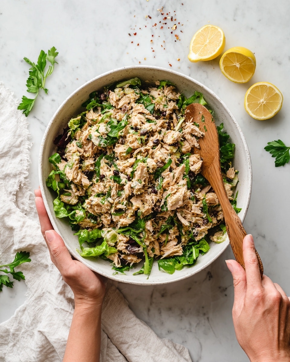 A large white bowl filled with a mixed salad made of shredded brown meat, beige textured chunks, and green leafy bits spread evenly throughout. A woman's hand is holding the bowl on the left side while another woman's hand stirs the mixture with a wooden spoon on the right. The bowl sits on a white marbled surface with two lemon wedges and some scattered green herbs and red seasoning nearby. A white cloth is partially visible under the bowl on the left side photo taken with an iphone --ar 4:5 --v 7