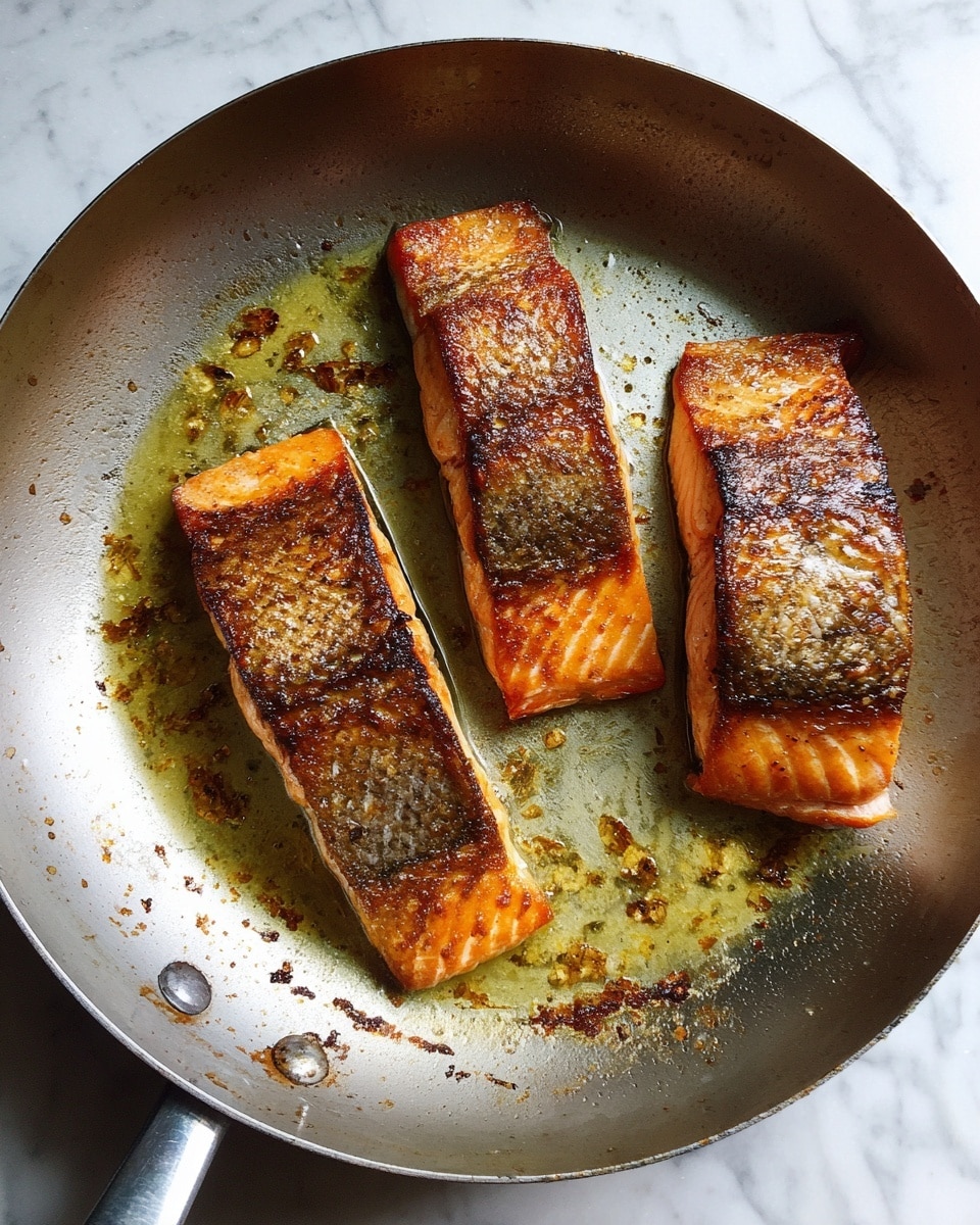 Three salmon fillets with a crispy, golden-brown surface are in a silver frying pan. The patties have a slightly shiny and textured look, showing the cooked skin side. The pan shows some oil and small browned bits around the fillets. The pan is placed on a white marbled surface. photo taken with an iphone --ar 4:5 --v 7
