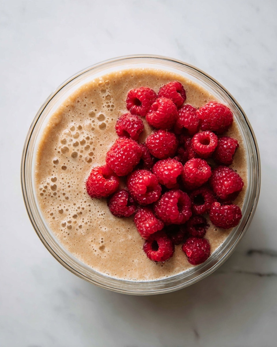 A clear glass bowl sits on a white marbled surface, filled with a thick, light brown mixture that has a slightly grainy texture. On top of the mixture, a pile of bright red raspberries is arranged on one side, bursting with juicy, round shapes. The rest of the surface shows the creamy, smooth mixture with small bubbles and a slightly uneven texture. The bowl's edges are visible but do not distract from the close-up focus on the raspberries and the mixture underneath. Photo taken with an iphone --ar 4:5 --v 7