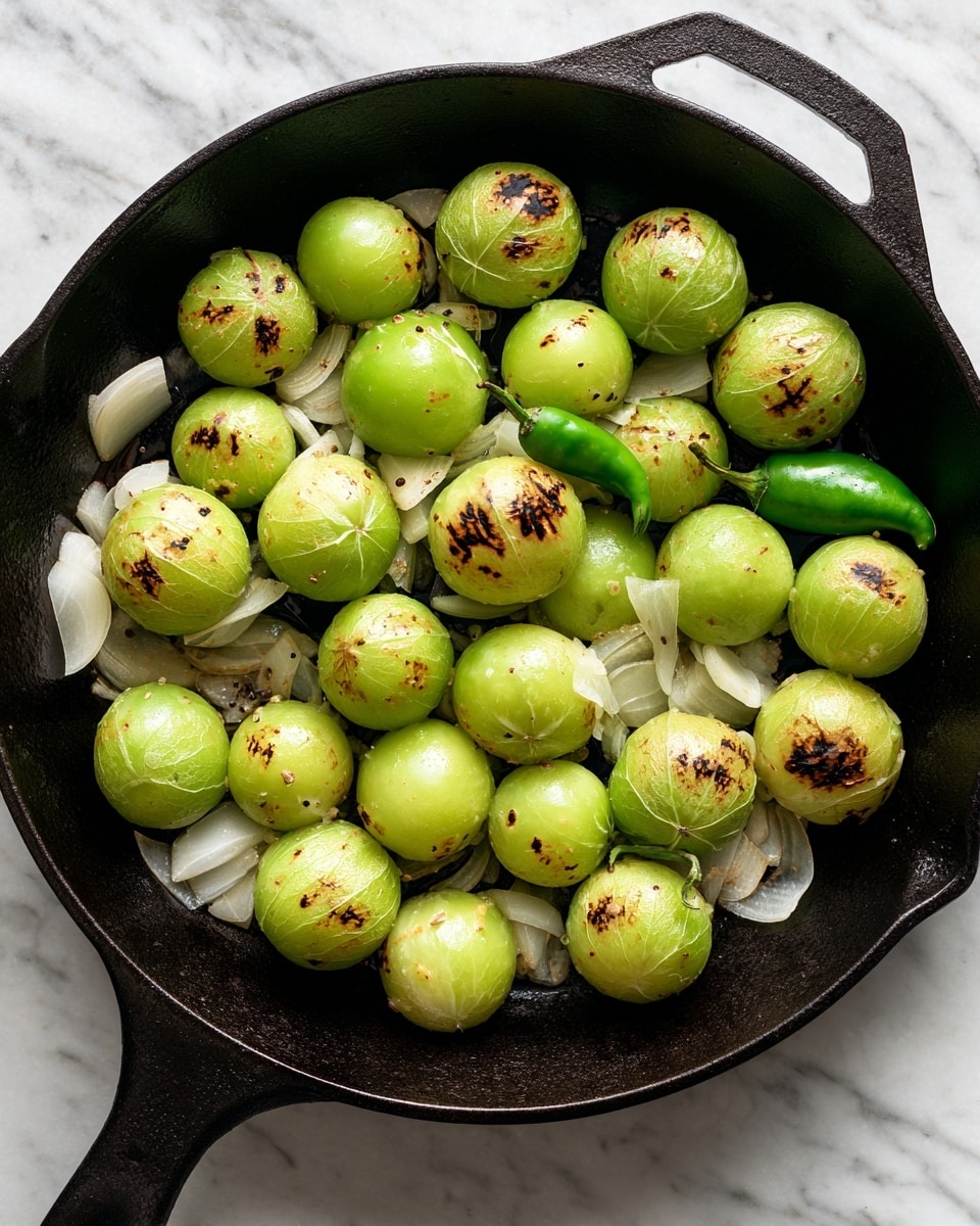 A black cast iron pan filled with round green tomatillos that have some light brown charred spots on their smooth skin. There are also several pieces of white onion slices scattered between the tomatillos, slightly cooked with soft edges. A single small green chili pepper lies in the pan, adding contrast to the mix. The pan rests on a white marbled surface. photo taken with an iphone --ar 4:5 --v 7