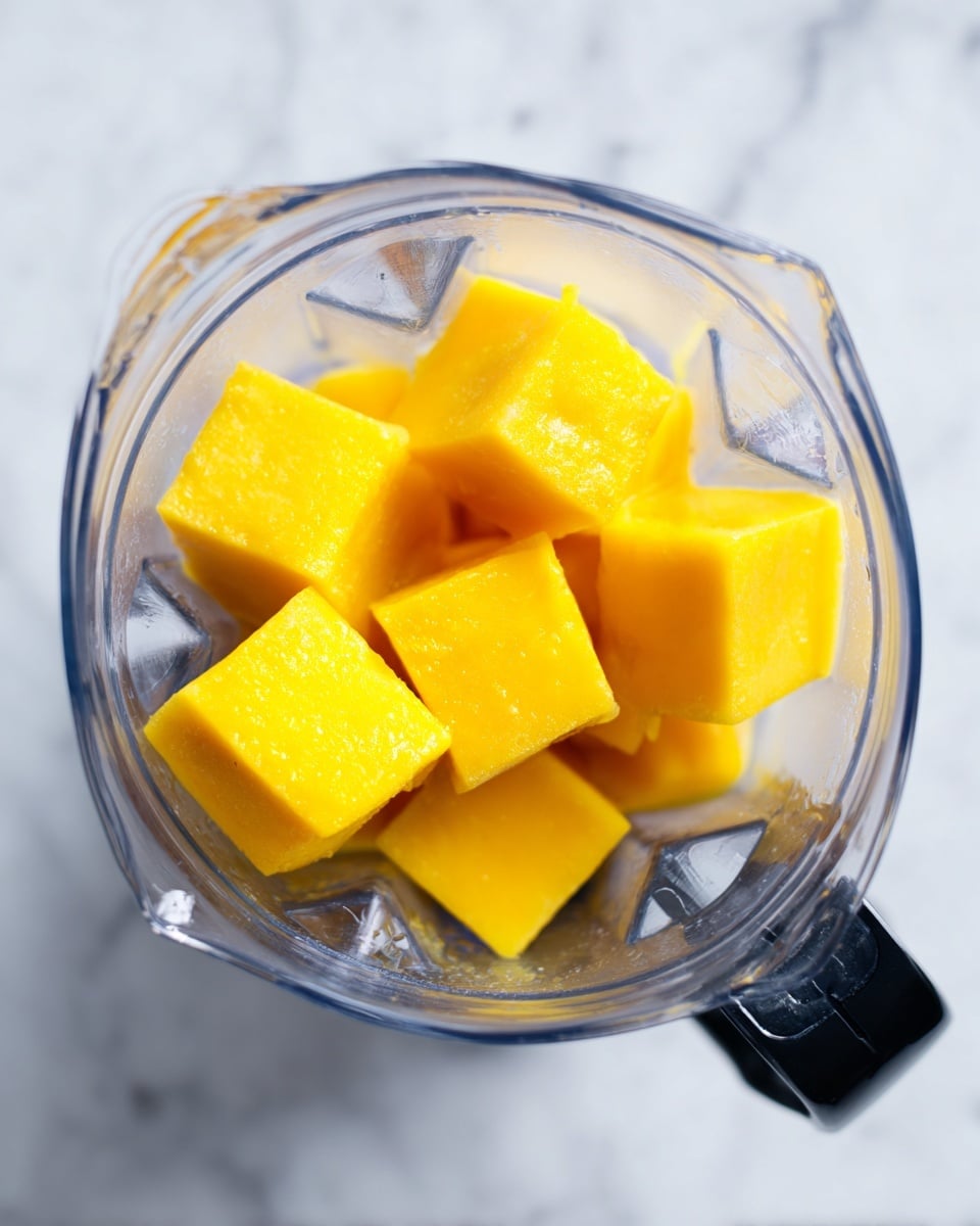The image shows a clear blender jar from a top view, containing several bright yellow frozen cubes stacked loosely inside. The cubes have a smooth, slightly frosty texture indicating they are frozen fruit or juice. The blender is placed on a white marbled surface, giving a clean and fresh look. The jar's clear sides and sharp angles are visible, with a black handle attached on the side. Photo taken with an iphone --ar 4:5 --v 7