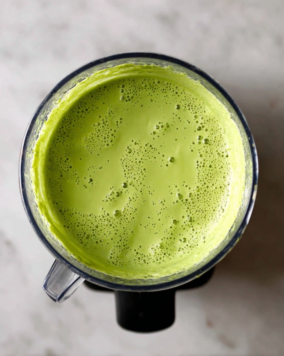 The image shows a close-up top view of a blender container filled with a smooth, bright green mixture. The mixture has a creamy texture with some small bubbles on the surface and tiny darker green specks evenly spread throughout, giving it a natural, fresh look. The inside walls of the blender container are coated with the green mixture, showing a thick consistency. The blender sits on a black base, and the background is a white marbled texture. photo taken with an iphone --ar 4:5 --v 7