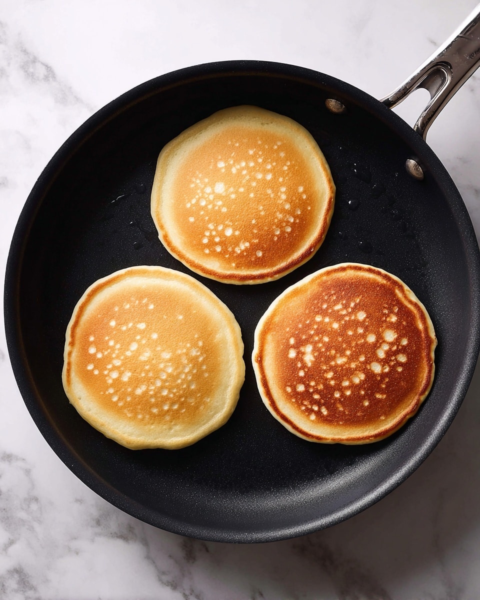 Three light brown pancake batter circles cook evenly on a black frying pan. Each pancake is smooth with small bubbles appearing on the surface, and they are spread out in a triangular shape in the pan. The pan has a matte black texture and is over a white marbled surface. The photo taken with an iphone --ar 4:5 --v 7