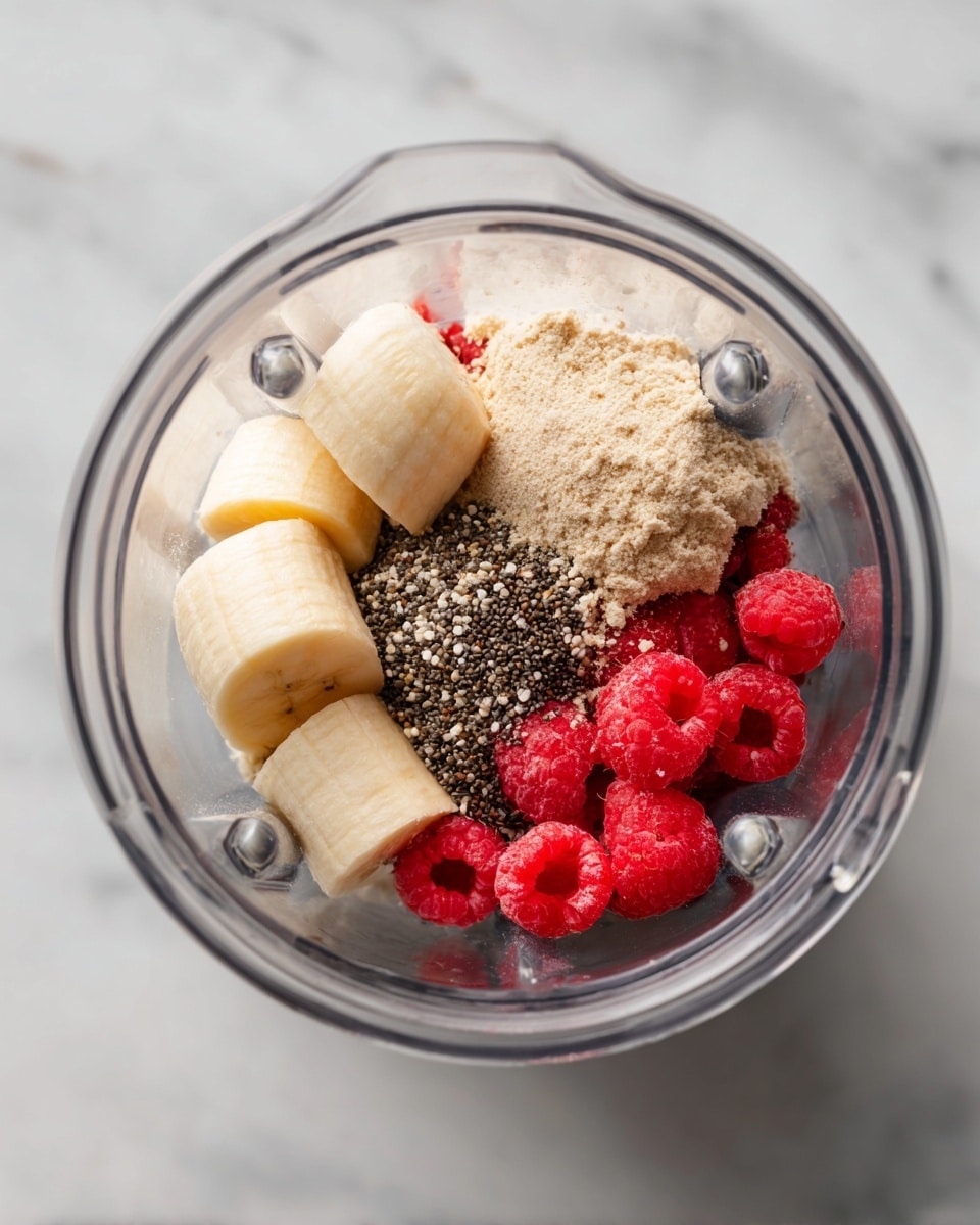 A clear blender jar sits on a white marbled surface, filled with four main ingredients in separate sections: two pale yellow banana halves, a pile of light beige protein powder with a crumbly texture, a small mound of tiny dark brown chia seeds, and a cluster of bright red raspberries. The ingredients are arranged side by side, showing a mix of smooth, powdery, and slightly textured surfaces. The blender jar is transparent, and the image is taken from above. photo taken with an iphone --ar 4:5 --v 7