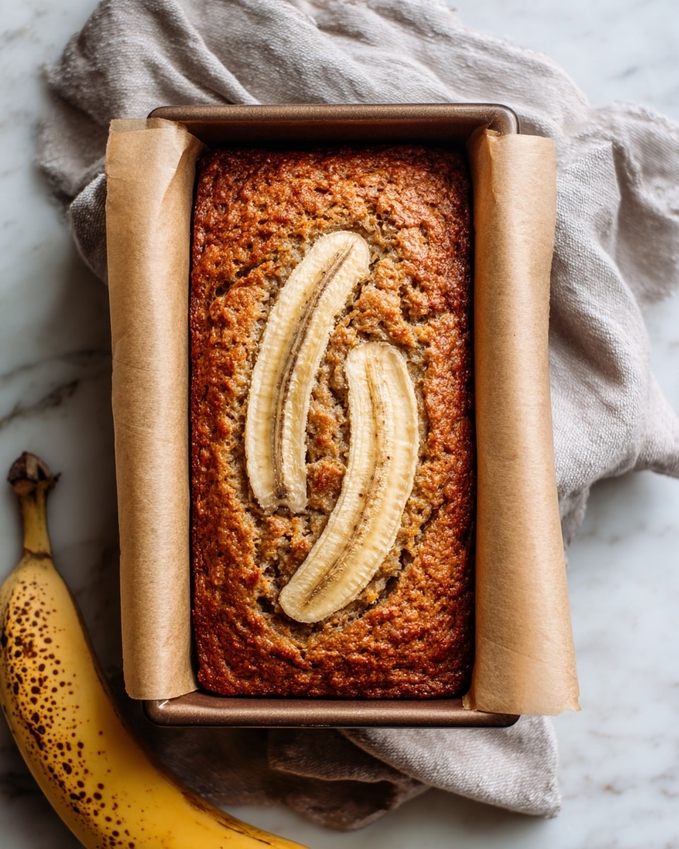 A loaf of banana bread with a textured, golden-brown crust sits in a rectangular brown baking pan lined with parchment paper, with two long banana halves placed on top center, showing their light beige color with dark seeds and a slightly glossy texture. The pan rests on a soft, light gray cloth against a white marbled background, with a ripe spotted banana positioned at the bottom left corner. The image is warm and inviting, emphasizing the homemade quality of the bread photo taken with an iphone --ar 4:5 --v 7