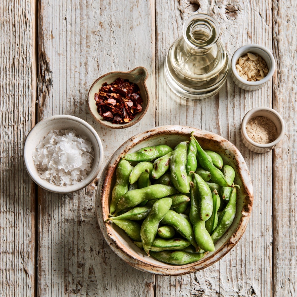 A white bowl filled with bright green edamame beans as the main layer, topped with a light sprinkle of coarse white salt and red chili flakes adding small hints of red and white across the beans, all on a white marbled surface; a small clear glass bowl with a light golden liquid is partially visible on the left side photo taken with an iphone --ar 4:5 --v 7