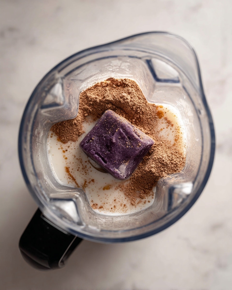 Inside a clear blender jar on a white marbled surface, there is a purple slab, likely a frozen fruit, laying flat in the center. Around the purple slab, there is a creamy white liquid filling the bottom, most likely milk, with light brown powder sprinkled on top of the purple slab and around it, creating a textured contrast. The blender jar has distinct sharp edges and a black lid attached to the top side. photo taken with an iphone --ar 4:5 --v 7