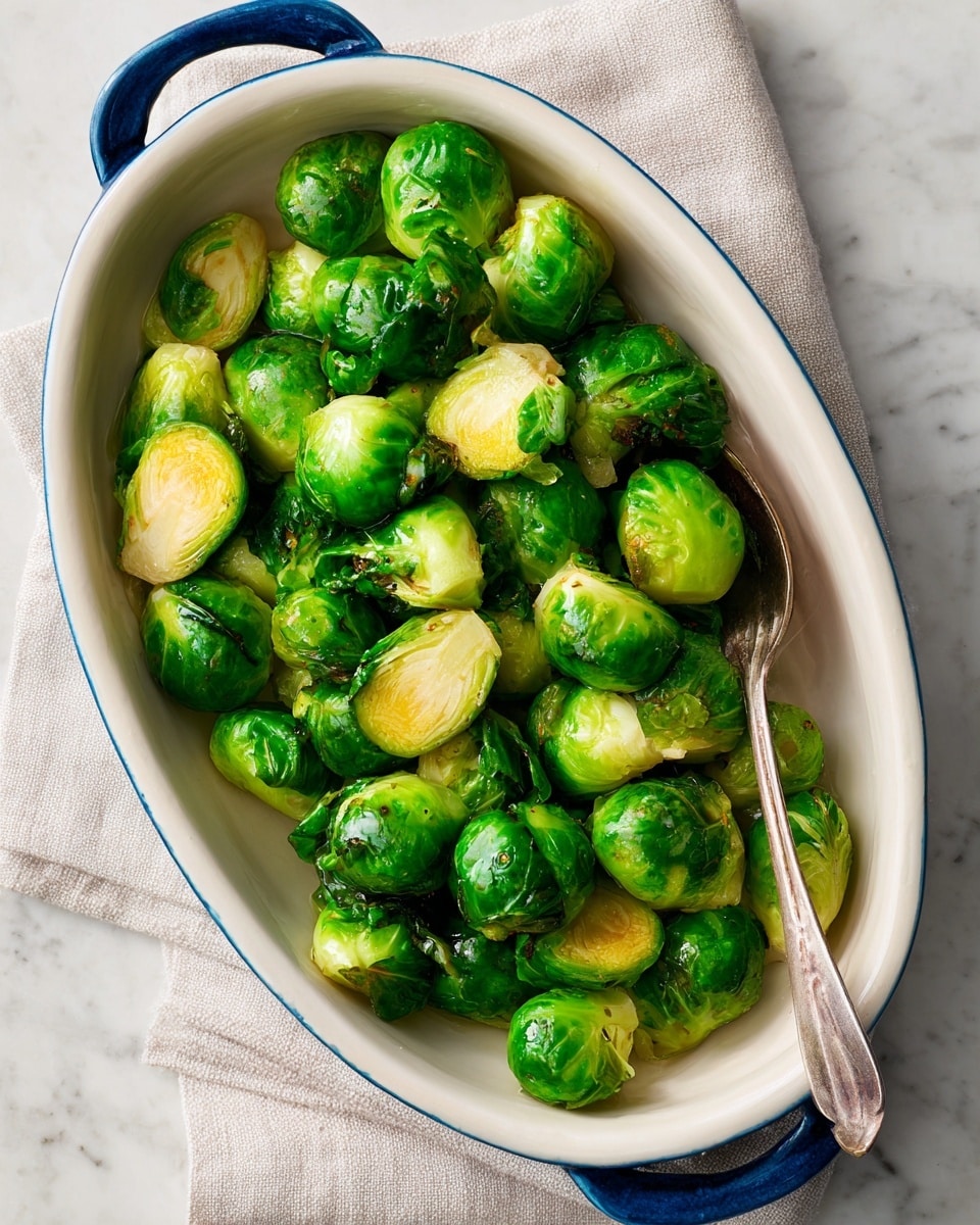 An oval white dish with blue handles holds one layer of cooked Brussels sprouts that are bright green with some darker green and slight browning from cooking, showing a mix of whole and halved pieces with a shiny, slightly oily texture. The dish sits on a white marbled surface. photo taken with an iphone --ar 4:5 --v 7