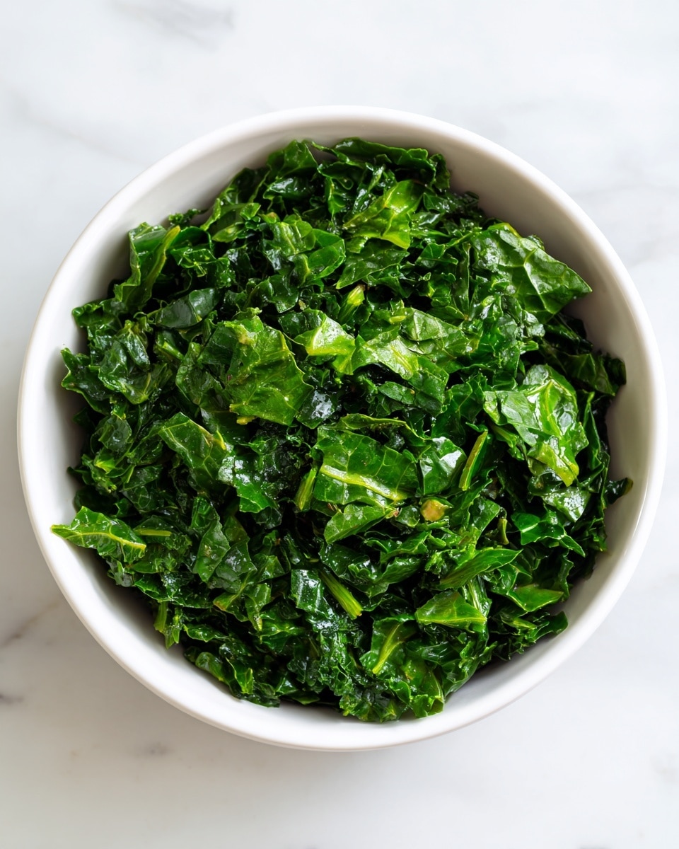 A white bowl filled with finely chopped dark green kale leaves that have a slightly wet, glossy texture, showing tiny bits of stems mixed evenly throughout. The bowl is placed on a white marbled surface, creating a clean and bright background that contrasts with the deep green color of the kale. photo taken with an iphone --ar 4:5 --v 7