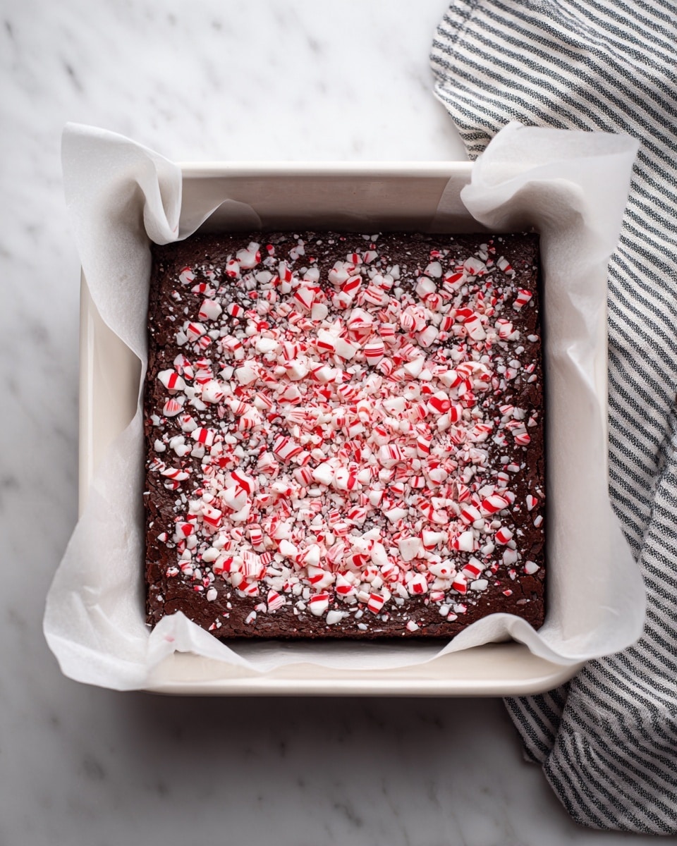 A square dark brown chocolate brownie sits inside a white square baking pan lined with white parchment paper that folds over the edges. The brownie is topped with a textured layer of crushed red and white peppermint candy pieces that cover the whole surface, adding a festive touch. The background is a clean white marbled surface with a black and white striped cloth partially visible on the right side. Photo taken with an iphone --ar 4:5 --v 7