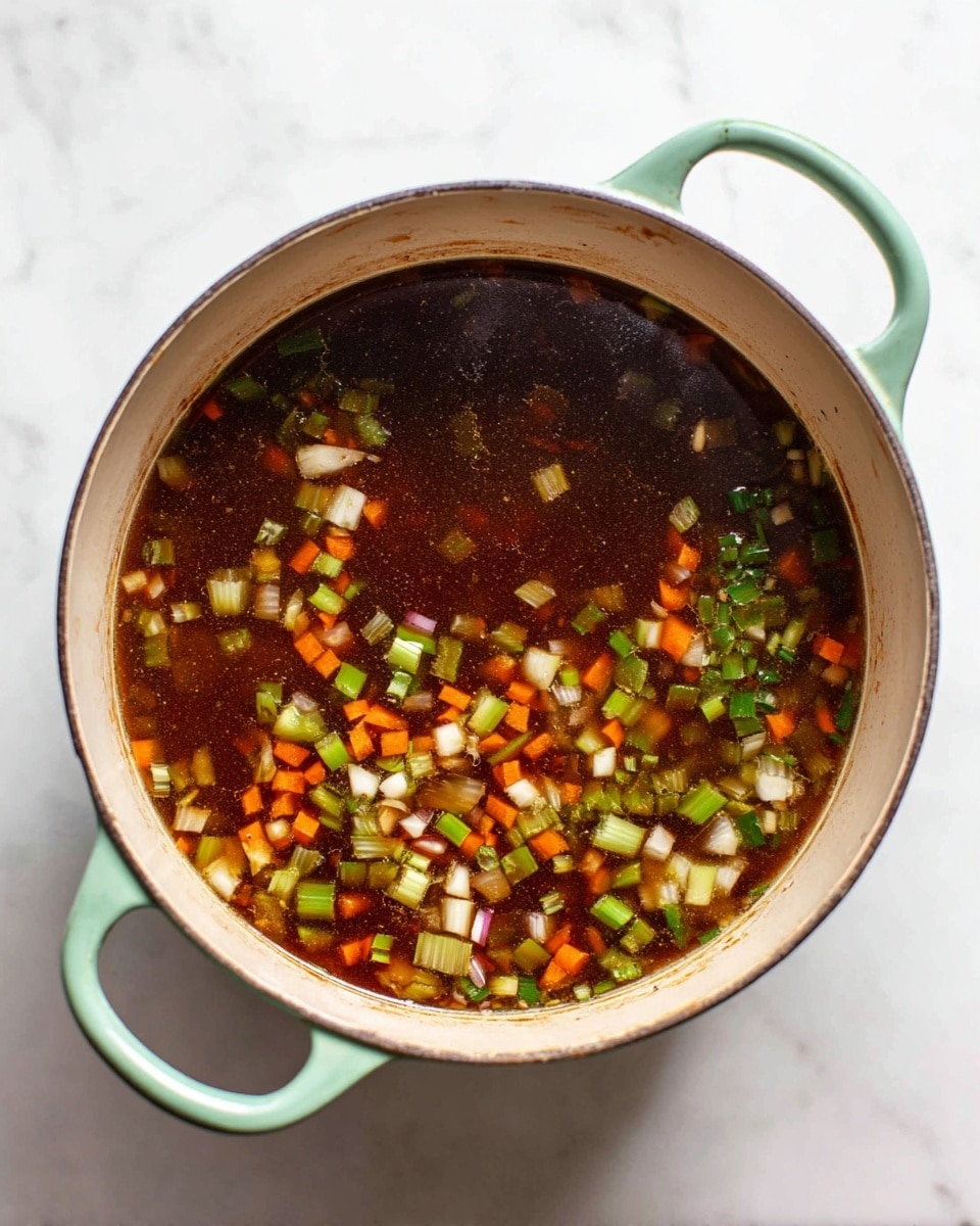 A round light green pot with two handles is filled with a dark brown broth that has visible oil spots on the surface. Floating in the broth are many small chopped pieces of light green celery, white garlic cloves, diced light purple onions, and small diced orange carrot cubes, all mixed evenly across the pot. The pot is placed on a white marbled surface. photo taken with an iphone --ar 4:5 --v 7
