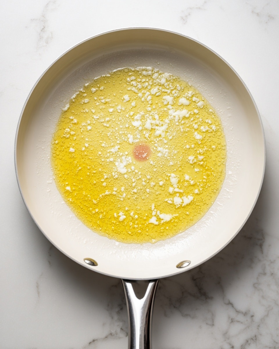 A white frying pan with a silver handle is filled with melted butter that spreads evenly across the surface. Small white clumps of butter solids float in the golden liquid, creating a textured pattern, especially near the center where a red circular heat indicator is visible. The edges of the pan are clean, showing the contrast between the white pan and the light yellow butter. The pan sits on a white marbled surface photo taken with an iphone --ar 4:5 --v 7