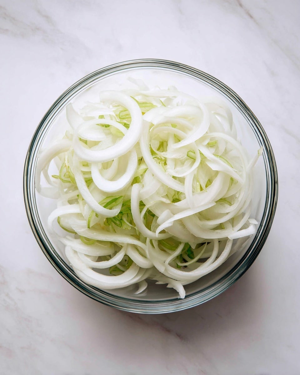 A clear glass bowl holds thinly sliced white onions with some light green parts visible near the edges. The onion slices are layered loosely and fill half of the bowl. The bowl is placed on a white marbled surface with soft natural light creating subtle shadows around it. photo taken with an iphone --ar 4:5 --v 7