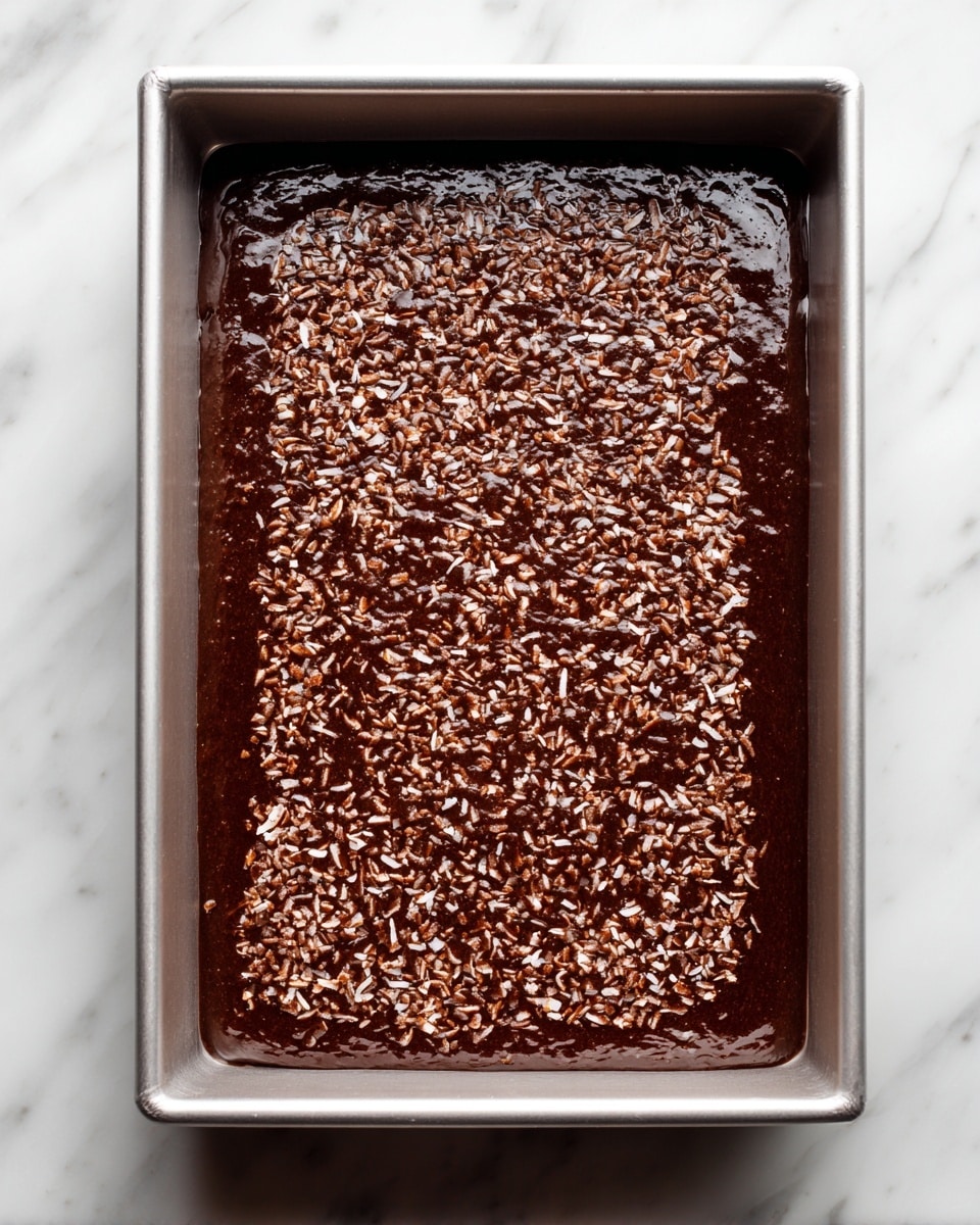 A rectangular metallic baking pan filled with one layer of thick, dark brown batter mixed with small light brown and white shredded pieces evenly spread inside. The pan sits on a white marbled surface with no other objects around. The batter has a glossy and slightly textured look, visible close to the edges of the pan. photo taken with an iphone --ar 4:5 --v 7