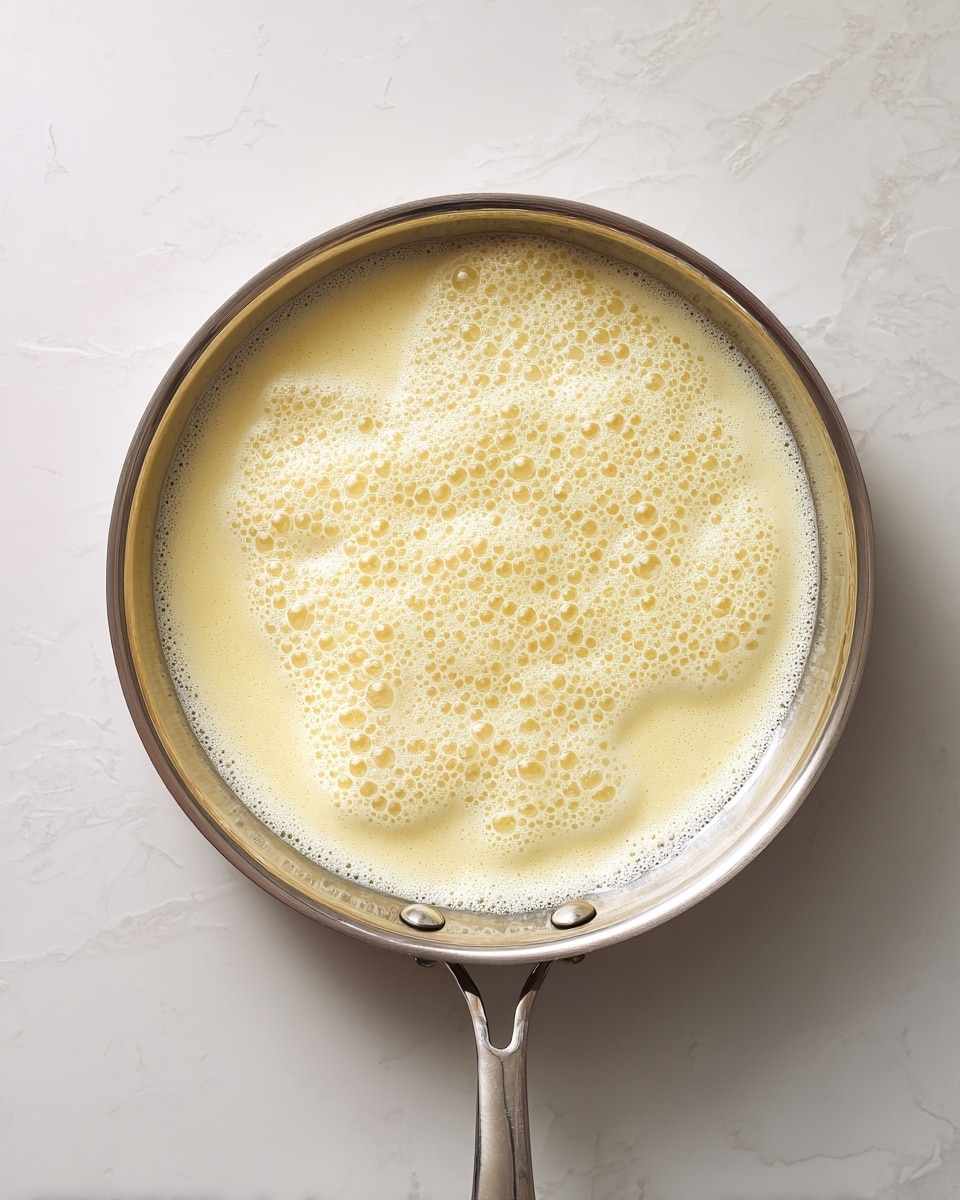 A top-down view of a metal pan filled with a foamy, light yellow liquid that has small bubbles spread evenly across the surface. The pan has a shiny, smooth texture with screws visible near the handle base. This scene is set on a white marbled textured surface that adds a soft contrast to the pan's silver tone. The liquid inside looks creamy and is spread evenly inside the pan, showing no additional layers or toppings photo taken with an iphone --ar 4:5 --v 7