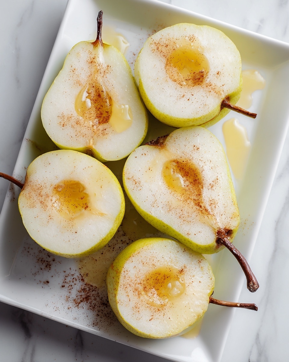 A white rectangular dish holds four pear halves arranged in two rows, each pear cut side up showing smooth, pale yellow flesh with a slight glossy texture. Each pear has a small, round hollow in the center filled with a dollop of melted butter or syrup, sprinkled generously with light brown cinnamon powder that also lightly dots the white dish surface. The pears have rough, thin greenish-brown skin around the edges, and one pear's stem is still attached. The dish rests on a white marbled surface, creating a clean and fresh background photo taken with an iphone --ar 4:5 --v 7