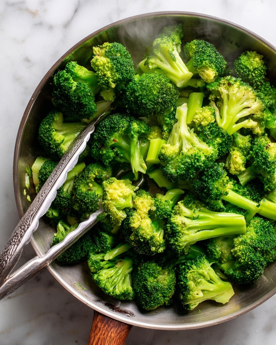 A shiny metal pan filled with bright green broccoli pieces styled in different sizes, with some showing their small, bumpy florets and others revealing smooth stalks. The broccoli is being held and mixed by a pair of silver tongs resting on the left side, with a slight steam rising around the vibrant vegetables. The pan sits on a white marbled surface that contrasts with the wooden texture around it, making the green color of the broccoli stand out clearly. The lighting highlights the freshness and texture of the broccoli. photo taken with an iphone --ar 4:5 --v 7