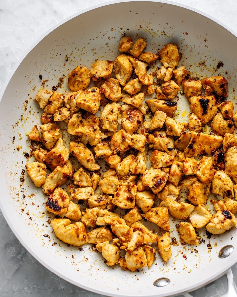 A white frying pan filled with many small pieces of cooked chicken that are golden brown and slightly charred, scattered all over the pan. The pan has some specks of seasoning and oil residue around the chicken, adding a textured look. The background is a white marbled surface. photo taken with an iphone --ar 4:5 --v 7