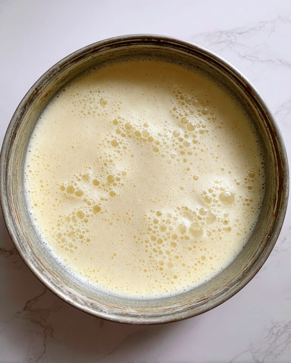 A large metal pan filled with a bubbling pale yellow liquid that has a foamy, frothy texture on top. The liquid's surface shows small bubbles and some thicker foam patches evenly spread across the pan. The pan is round with a slightly worn rim visible at the top edge, placed on a white marbled surface photo taken with an iphone --ar 4:5 --v 7