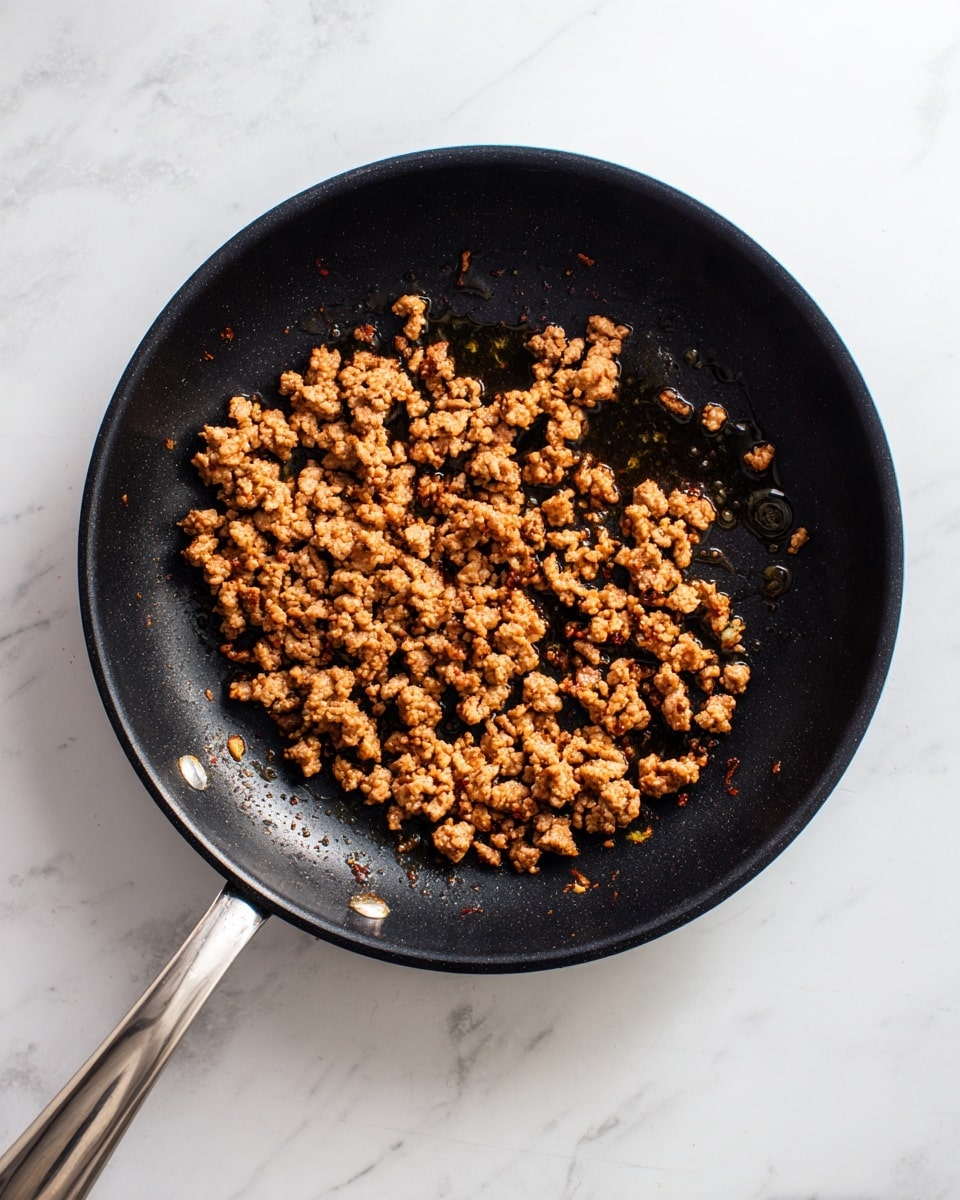 In a black frying pan, there is one layer of cooked ground meat scattered evenly but not packed tightly. The meat pieces vary in size, showing some with brown, slightly crispy edges and others softer and lighter brown in color. Small pools of cooking oil add a shiny texture around the meat bits. The pan handle is metallic and extends out to the left side. The pan is placed on a white marbled surface with soft shadows falling on it. photo taken with an iphone --ar 4:5 --v 7