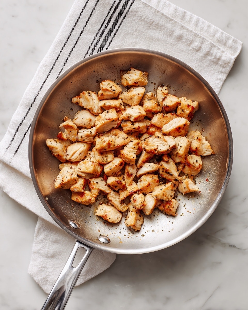A silver frying pan holds small pieces of cooked chicken that are light golden brown with some darker spots from cooking. The chicken pieces are uneven in size and spread mainly in the center and bottom right of the pan. The pan is placed on a white marbled surface with a white towel with black stripes folded and lying on the top left corner. The pan’s handle extends out towards the bottom left corner of the image. photo taken with an iphone --ar 4:5 --v 7