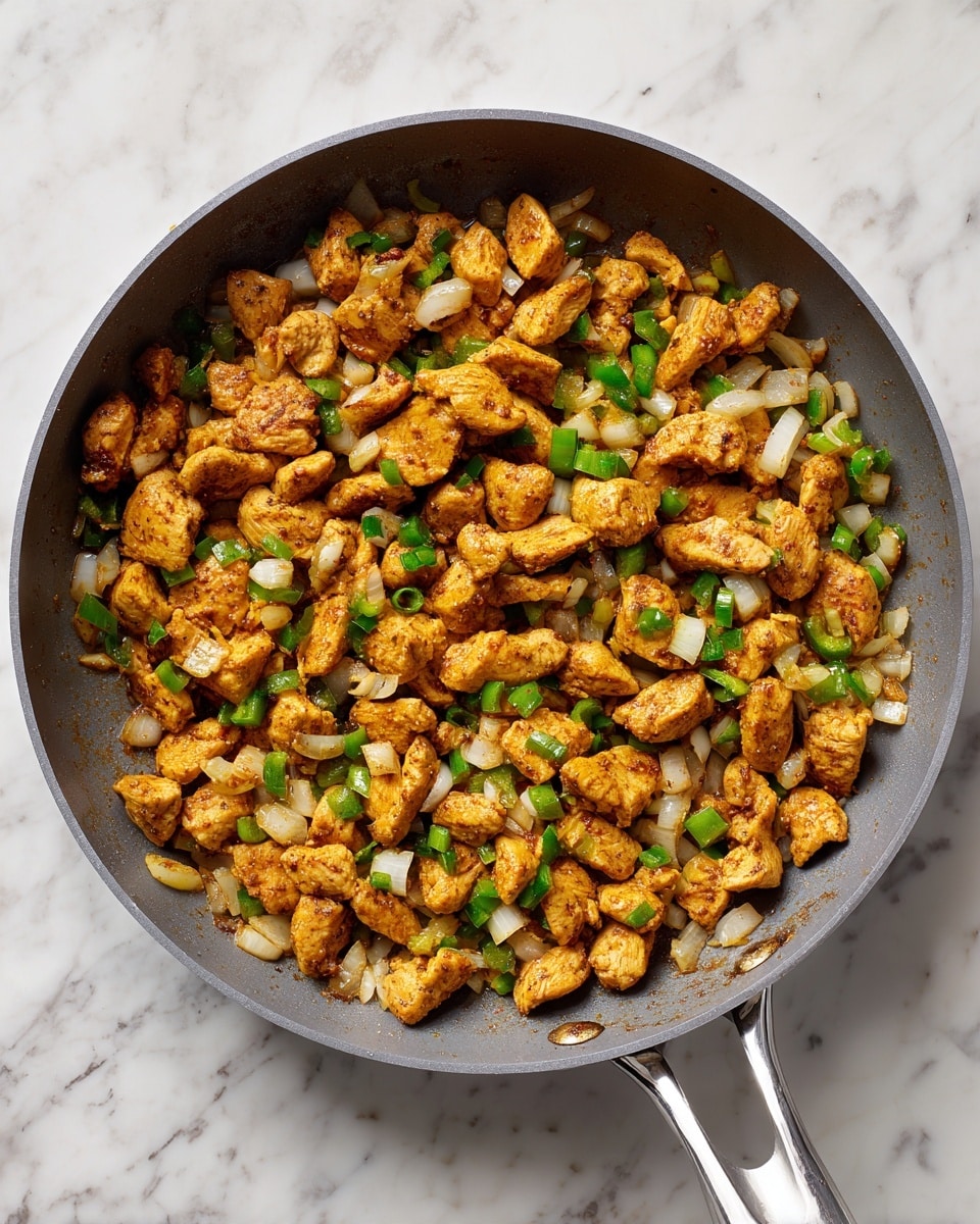 A large grey pan with a silver handle is filled with cooked small pieces of golden-brown chicken mixed with diced white onions and green bell peppers, all spread evenly across the pan. The pan sits on a white marbled surface. The cooked chicken pieces look tender with a light brown spice coating, and the onions and peppers add touches of white and green color throughout. photo taken with an iphone --ar 4:5 --v 7
