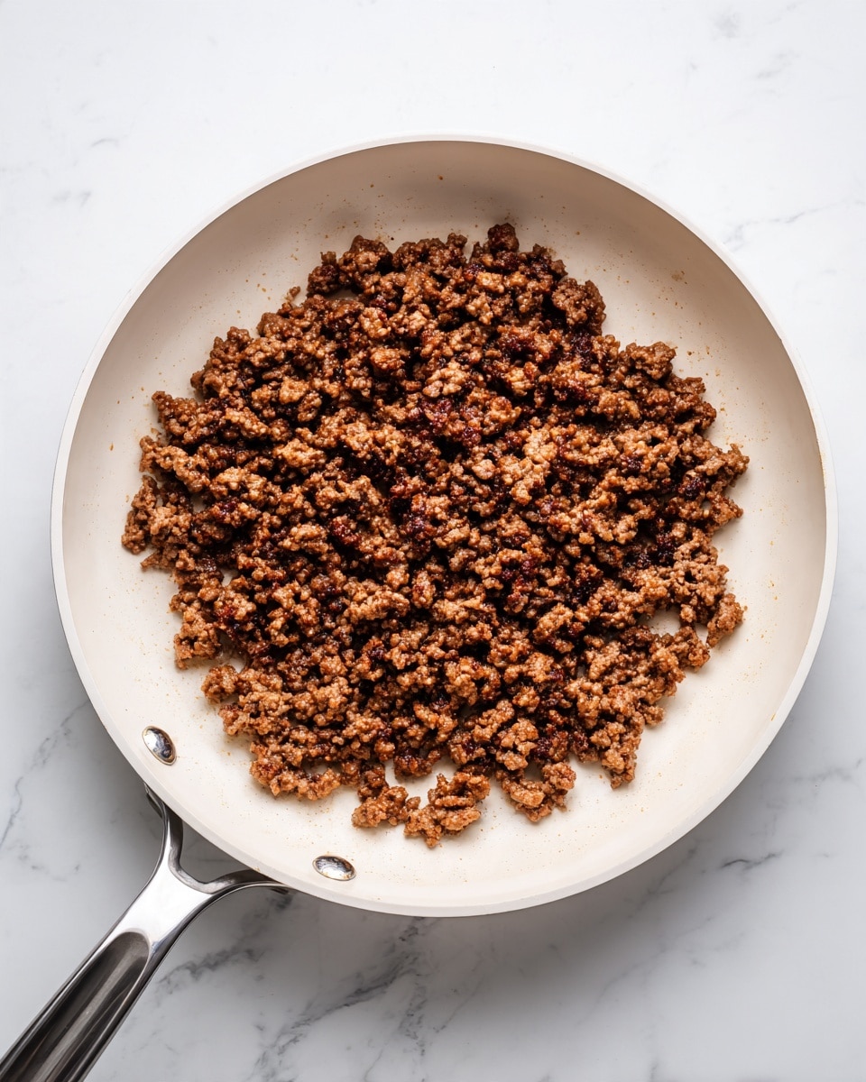 In a large white frying pan, there are many small pieces of browned ground meat spread evenly across the surface. The meat pieces have a mix of dark brown and lighter brown shades, showing they are cooked with a slightly crisp texture on the outside. The pan itself has a smooth, clean white inside and a silver handle that extends off to the right. The pan is placed on a white marbled surface that provides a lighter, clean background with subtle gray veins. photo taken with an iphone --ar 4:5 --v 7