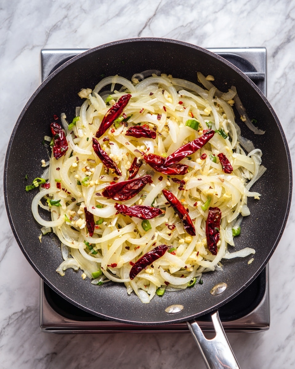 A black non-stick frying pan holds a mix of light yellow and white sliced onions spread in the pan, with several small dried red chili peppers scattered evenly on top. There are also some small green pieces resembling sliced green chilies and minced garlic sprinkled throughout the onions. The pan sits on a stove with a white marbled surface background. photo taken with an iphone --ar 4:5 --v 7