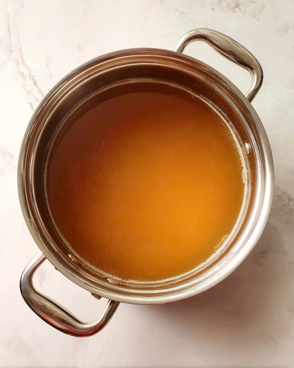 A shiny stainless steel pot with two handles is filled halfway with light brown clear broth. The pot sits on a white marbled surface, with the steam faintly rising from the warm liquid inside. The broth is smooth and even in color, showing no other solid pieces or layers. Photo taken with an iphone --ar 4:5 --v 7
