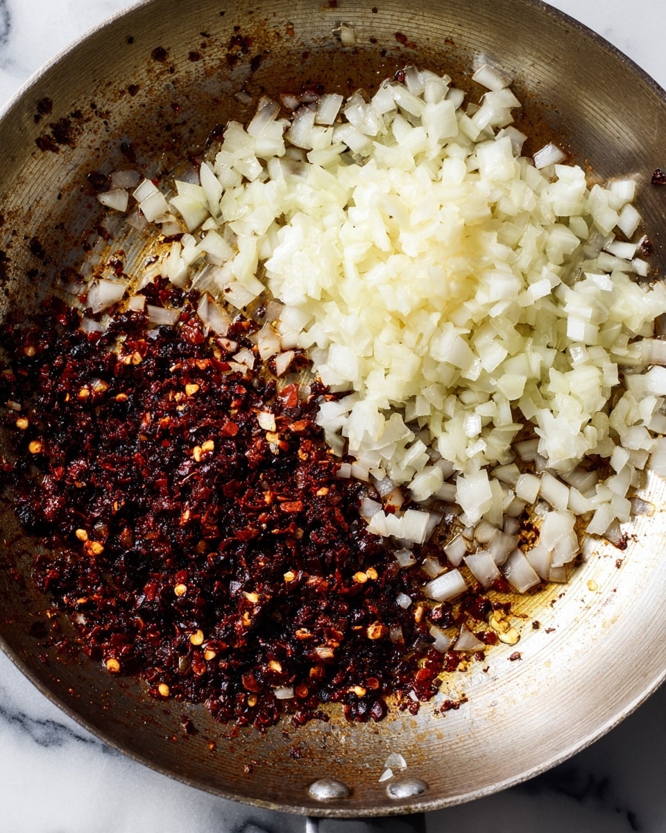 A close-up view of a pan showing two main layers: on one side, small white chopped onion pieces with a fresh, slightly shiny texture, and on the other side, coarse black chili flakes mixed with oil, creating a dark, oily layer with a slightly shiny surface. The pan has a metal surface with some browning and oil stains around the edges. The background is a white marbled texture. photo taken with an iphone --ar 4:5 --v 7